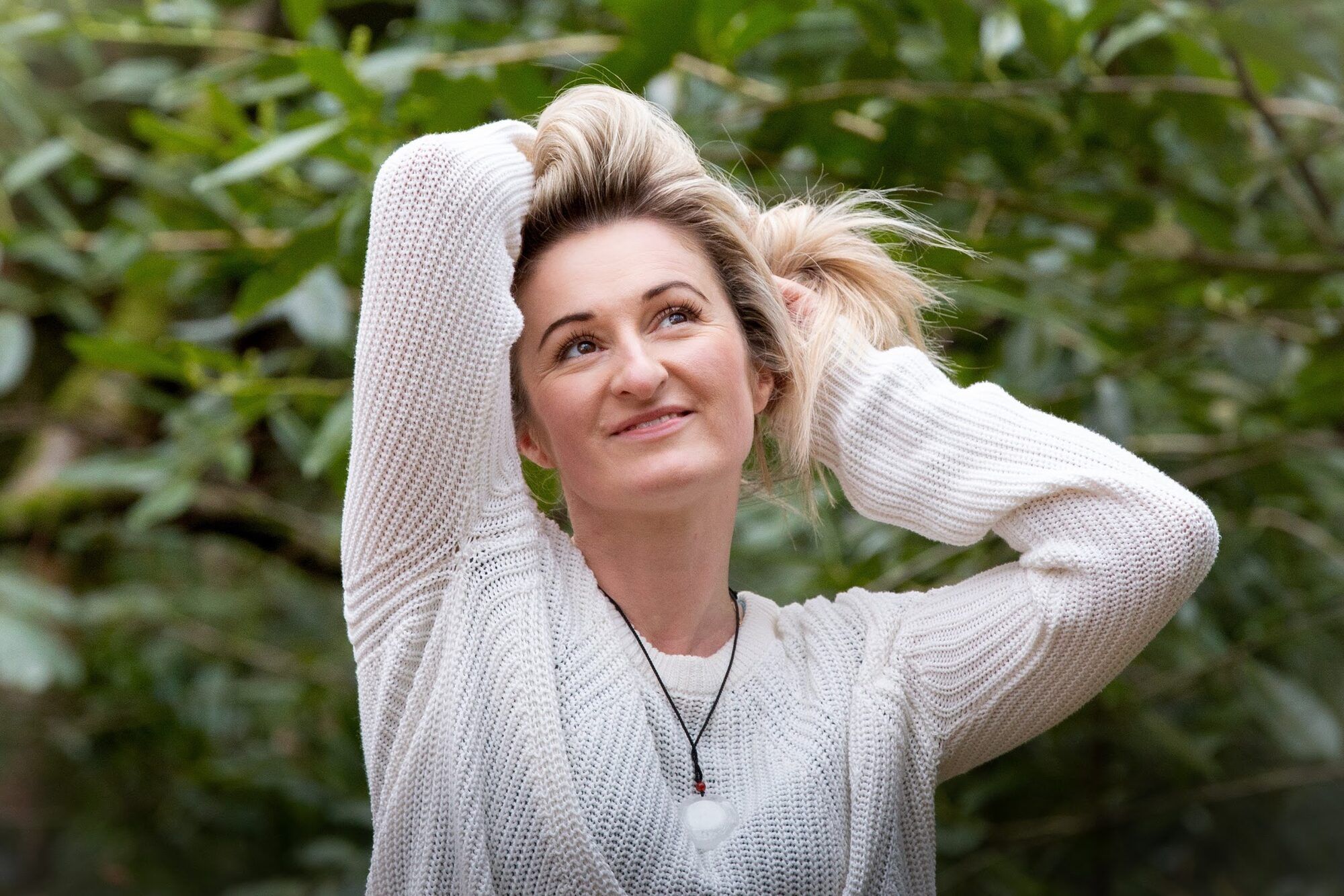 Blonde woman in a cream jumper, hands in her hair, greenery behind