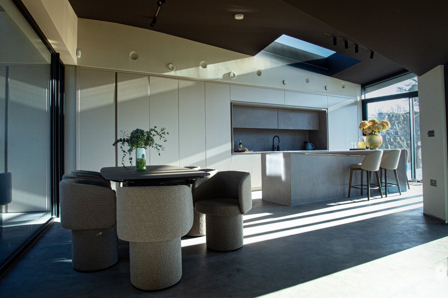 Open-plan kitchen and dining area with a grey island, rounded upholstered chairs and sunlight striping the dark floor